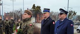 Żołnierz trzyma wieniec za nim stoją obok siebie funkcjonariusz Państwowej Straży Pożarnej oraz Policji.