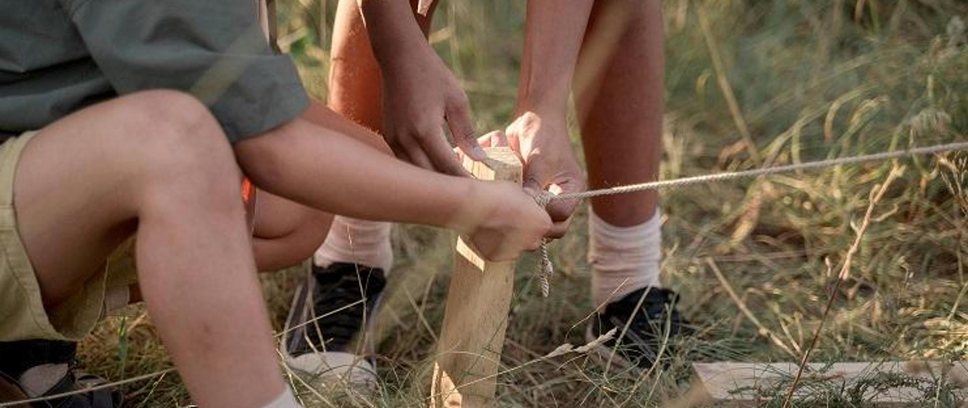 na zdjęciu widać ręce dwóch chłopców, prawdopodobnie harcerzy, którzy kucają i rozkładają namiot na polanie