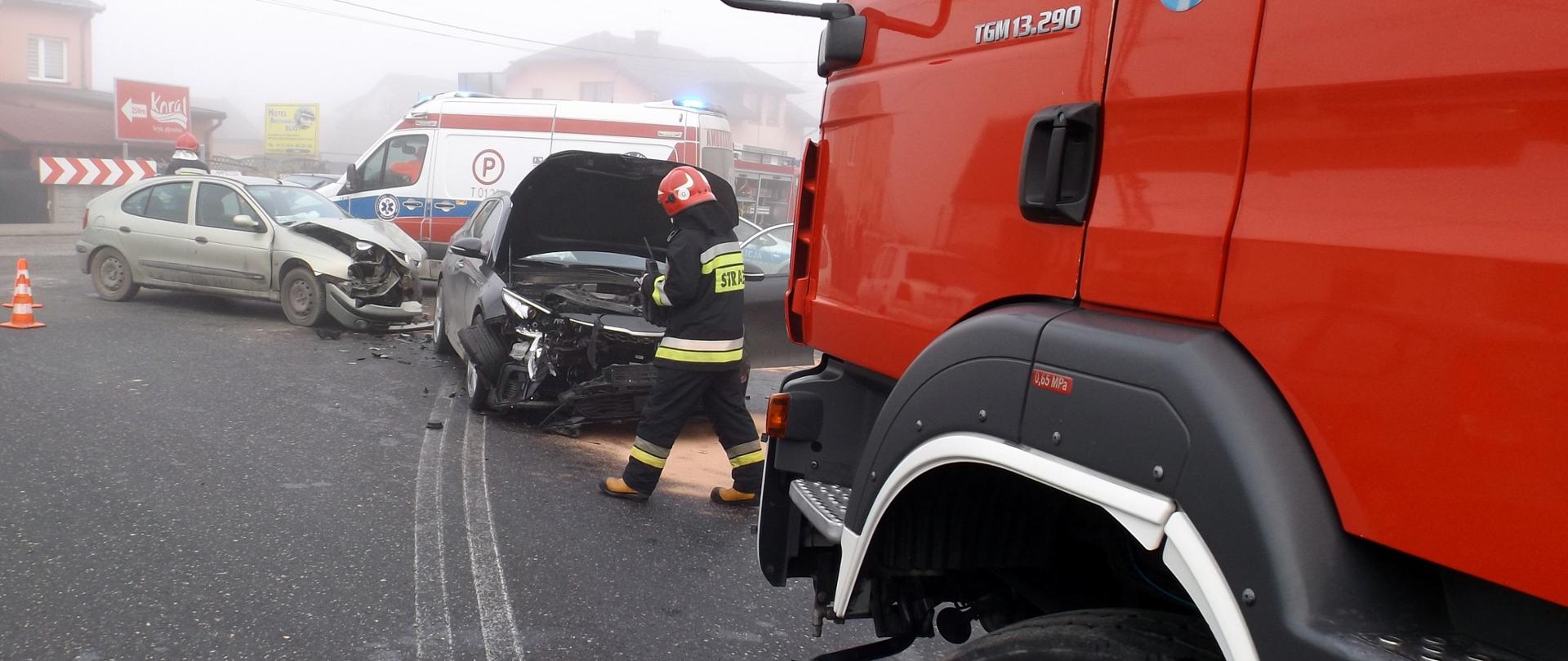 Na pierwszym planie znajduje się samochód strażacki, za nim strażak z radiotelefonem w reku oraz rozbite samochody osobowe: czarna Kia oraz srebrny Renault. Samochody mają rozbite przody. W tle stoi Karetka Pogotowia Ratunkowego