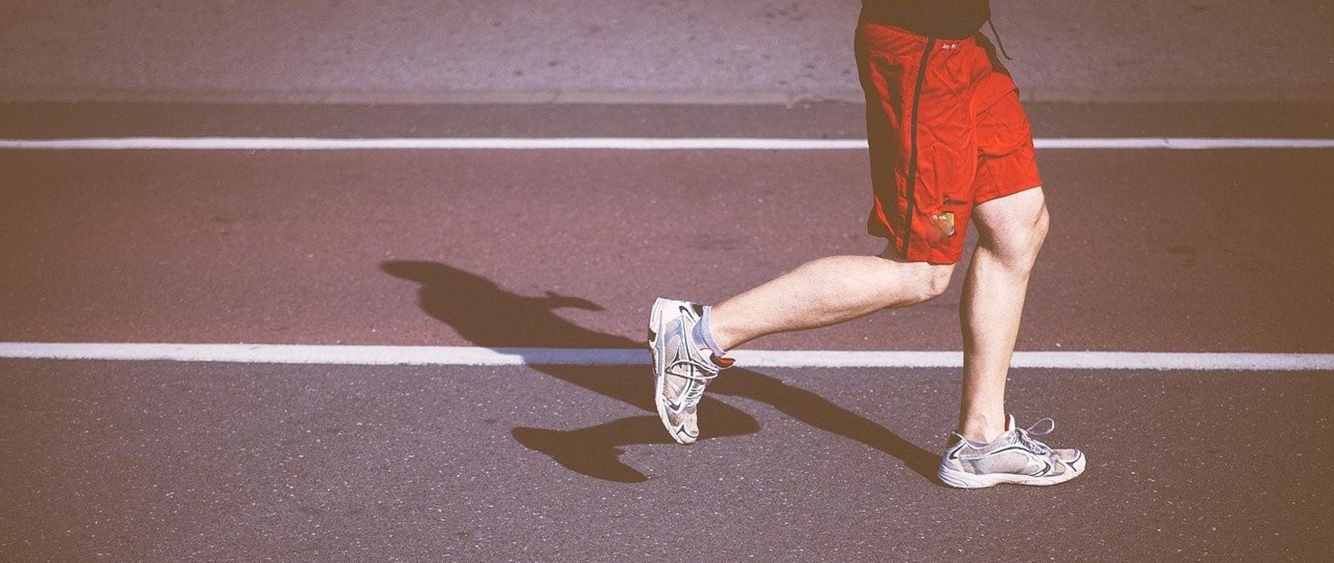 Grafika przedstawia sylwetkę człowieka od pasa w dół w trakcie biegu na bieżni, ubranego w czerwone krótkie spodenki i szare buty sportowe.