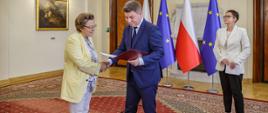 Minister Jan Grabiec wręcza akt powołania do Rady KSAP prof. Małgorzacie Bonikowskiej, obok stoi szefowa służby cywilnej Anita Noskowska-Piątkowska; w tle flagi Polski i Unii Europejskiej