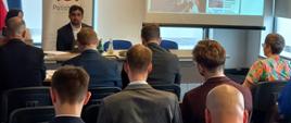 Przy stole konferencyjnym stojącym obok banneru i ekranu z prezentacją siedzi mężczyzna i przemawia do uczestników spotkania