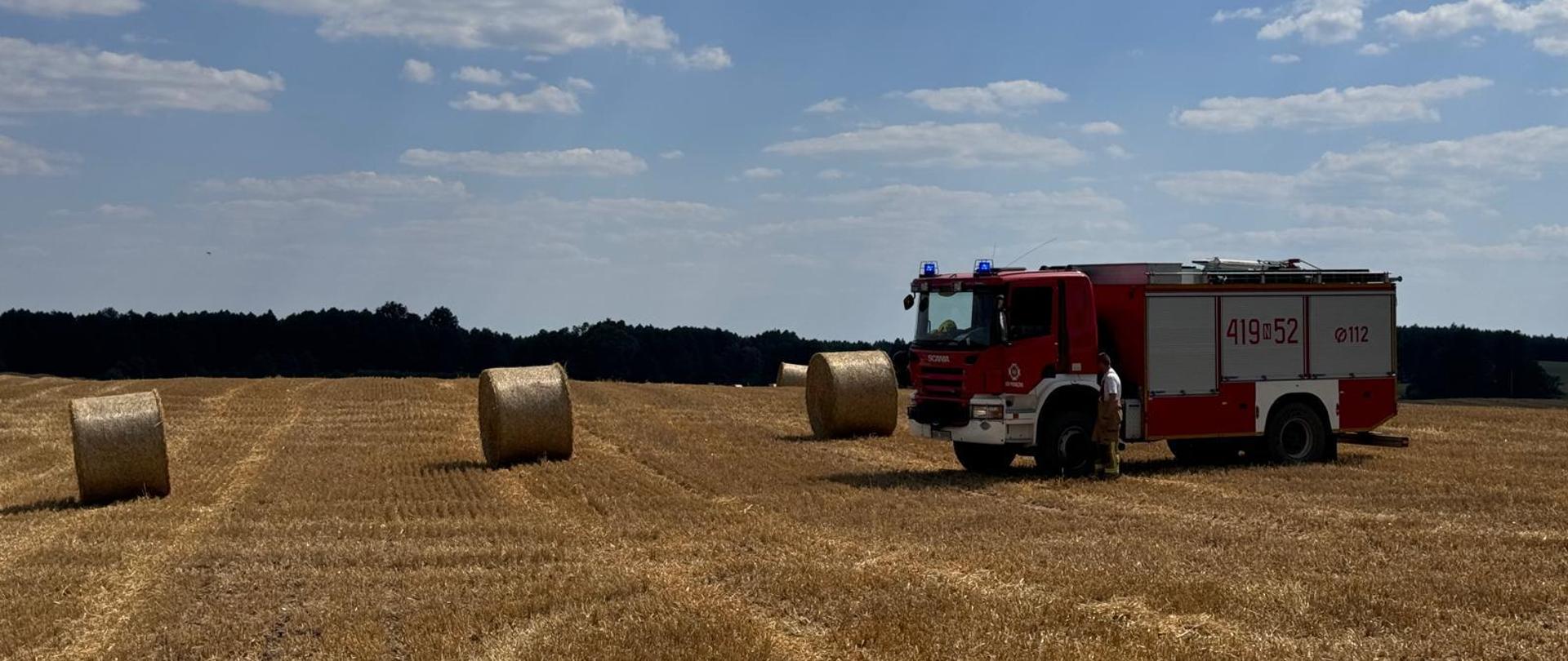 Na ściernisku stoi duży samochód strażacki. Na polu i przy samochodzie baloty słomy. Niebieskie niebo, słonecznie.