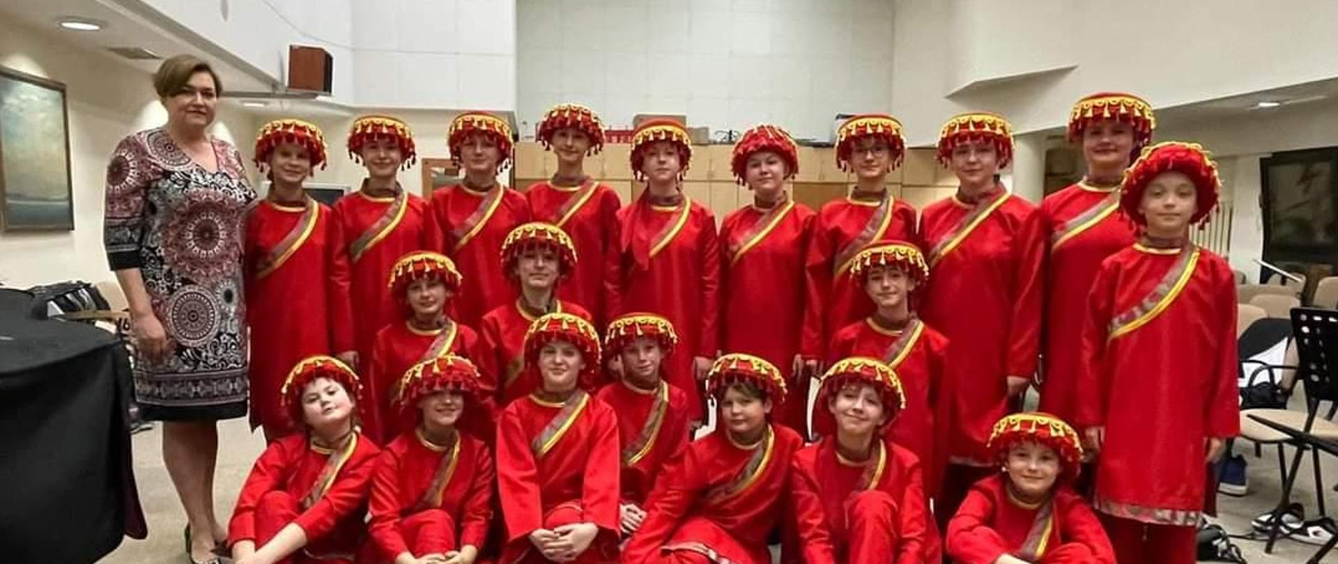 na białym tle stoją ustawione obok siebie dzieci w czerwonych strojach z czerwonymi nakryciami głowy i żóltymi fredzlami na nich