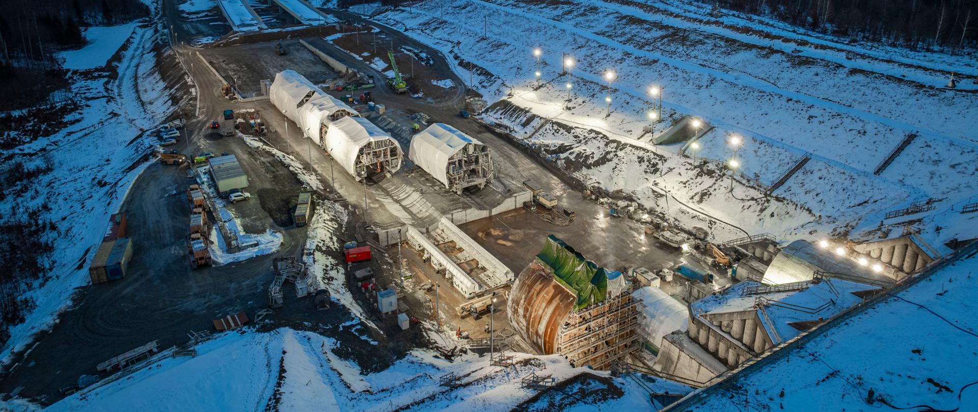 Zdjęcie wykonane z lotu ptaka pokazuje duży, oświetlony plac budowy pośród zaśnieżonego terenu. Widać kilka ogromnych, cylindrycznych elementów konstrukcyjnych ułożonych w rzędzie oraz maszyny budowlane. Po bokach rozciągają się wykopy i drogi dojazdowe pokryte śniegiem. W tle znajduje się ciemny las i rozległy, zimowy krajobraz sięgający aż po horyzont. Cała budowa jasnym światłem kontrastuje z otaczającą ją ciemnością wieczoru.