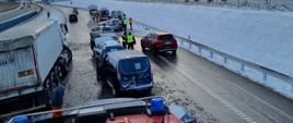Zdjęcie obrazuje drogę ekspresową nr 7 i zrobione jest z dachu pojazdu ratowniczo-gaśniczego. Na drodze znajdują się uszkodzone pojazdy osobowe, blokujące częściowo dwa pasy ruchu. Drugi samochód biorący udział w zdarzeniu znajduje się na pasie zieleni. Obok uszkodzonych samochodów znajduje się także ciężarówka, która ugrzęzłą na poboczu drogi. Na zdjęciu widnieją także strażacy w umundurowaniu specjalnym i policjanci.