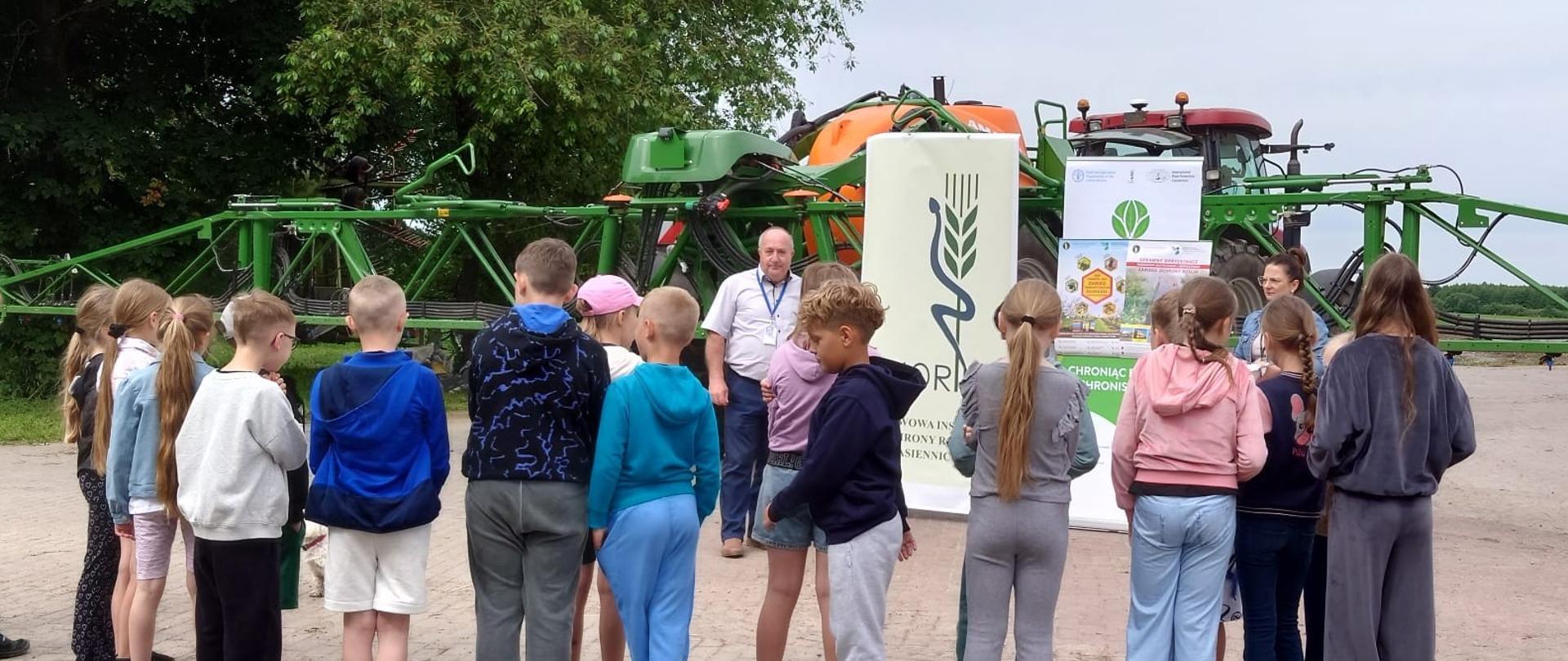Grupa dzieci stoi na placu i uczestniczy w pokazie dużego opryskiwacza rolniczego. Przed maszyną znajdują się osoby prowadzące prezentację oraz tablice informacyjne związane z ochroną roślin. W tle widoczne są drzewa i pochmurne niebo.