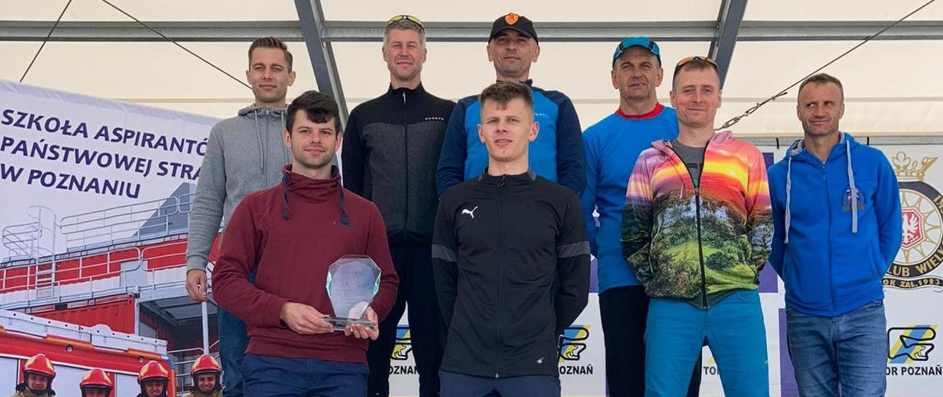 Na zdjęciu widać kilku mężczyzn w ubraniu sportowym i cywilnym znajdujących się na scenie oraz podeście podczas wręczania nagród w zawodach biegowych. Panowie to funkcjonariusze Państwowej Straży Pożarnej. 