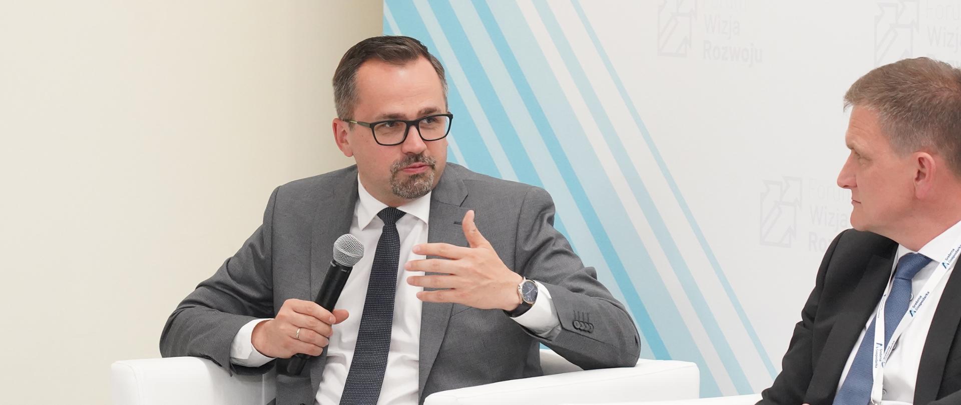 Dwie osoby rozmawiają, siedząc obok siebie w białych fotelach. Pierwszy z lewej siedzi wiceminister Marcin Horała.