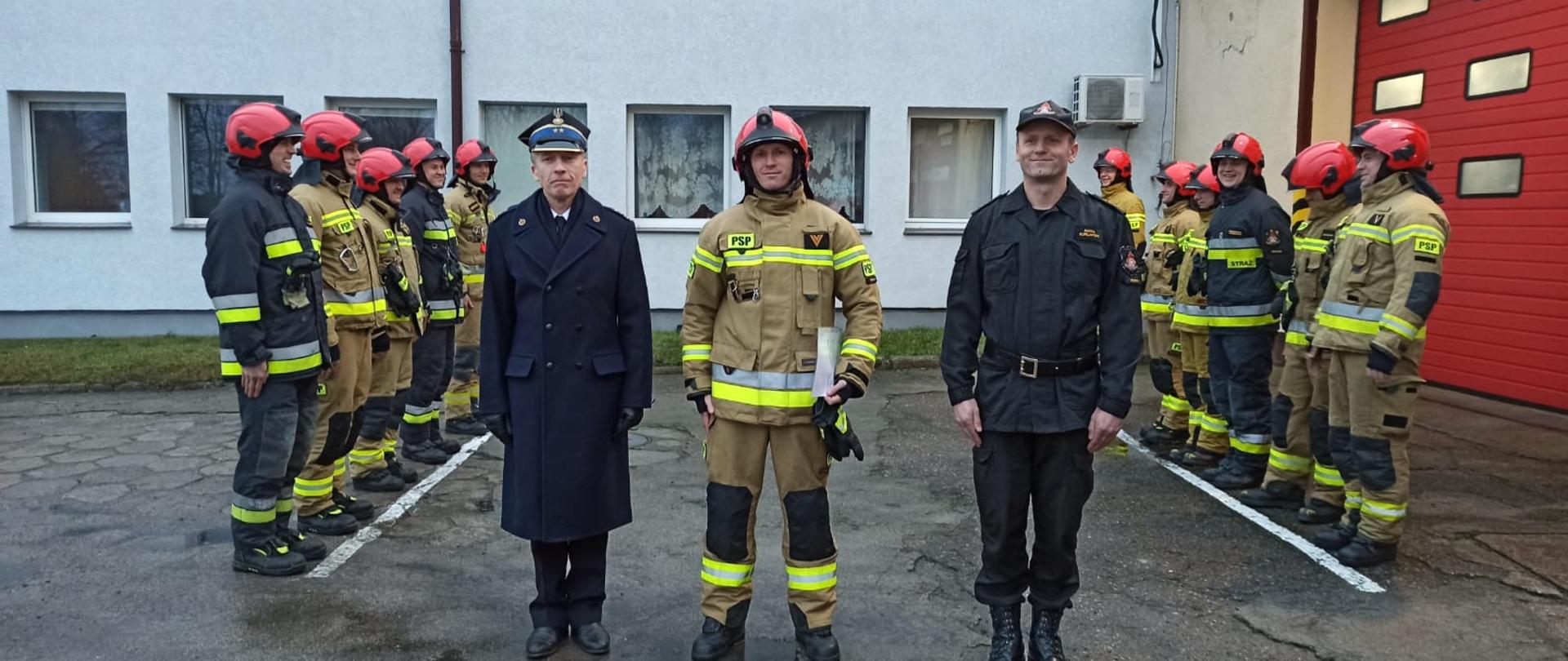 Zdjęcie przedstawia plac przed czerwonymi bramami garażowymi Jednostki Ratowniczo-Gaśniczej w Świdwinie. Przed bramą stoi zmiana służbowa w piaskowych ubraniach specjalnych z czerwonymi hełmami - stoją wzdłuż namalowanej białej linii. Na pierwszym planie znajduje się Komendant Powiatowy PSP w Świdwinie, dowódca Jednostki ratowniczo-Gaśniczej w Świdwinie ubrani w czarny mundur czytając coś z dokumentów. Obok niego stoi wyróżniony strażak ubrany w piaskowy mundur i czerwony hełm, trzymający w ręku dokumenty. 