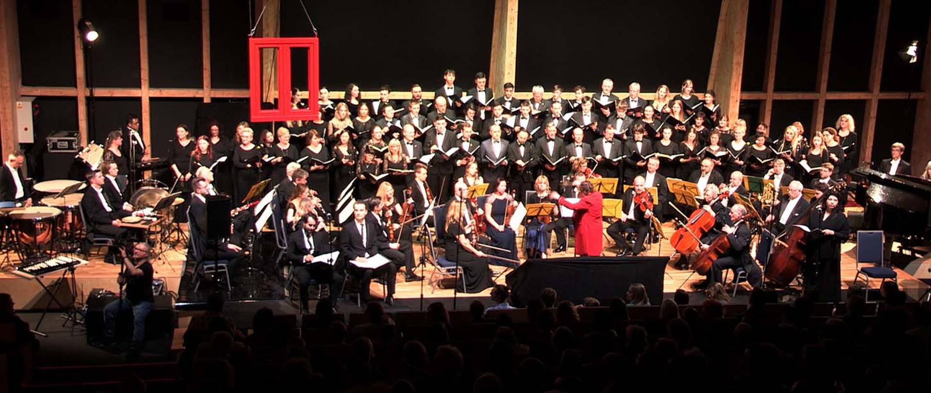 Zdjęcie sceny na której w tylnej części stoi chór składający się z kilkudziesięciu osób, z przodu i po bokach liczna orkiestra symfoniczna, przed nimi na podwyższeniu stoi dyrygentka.