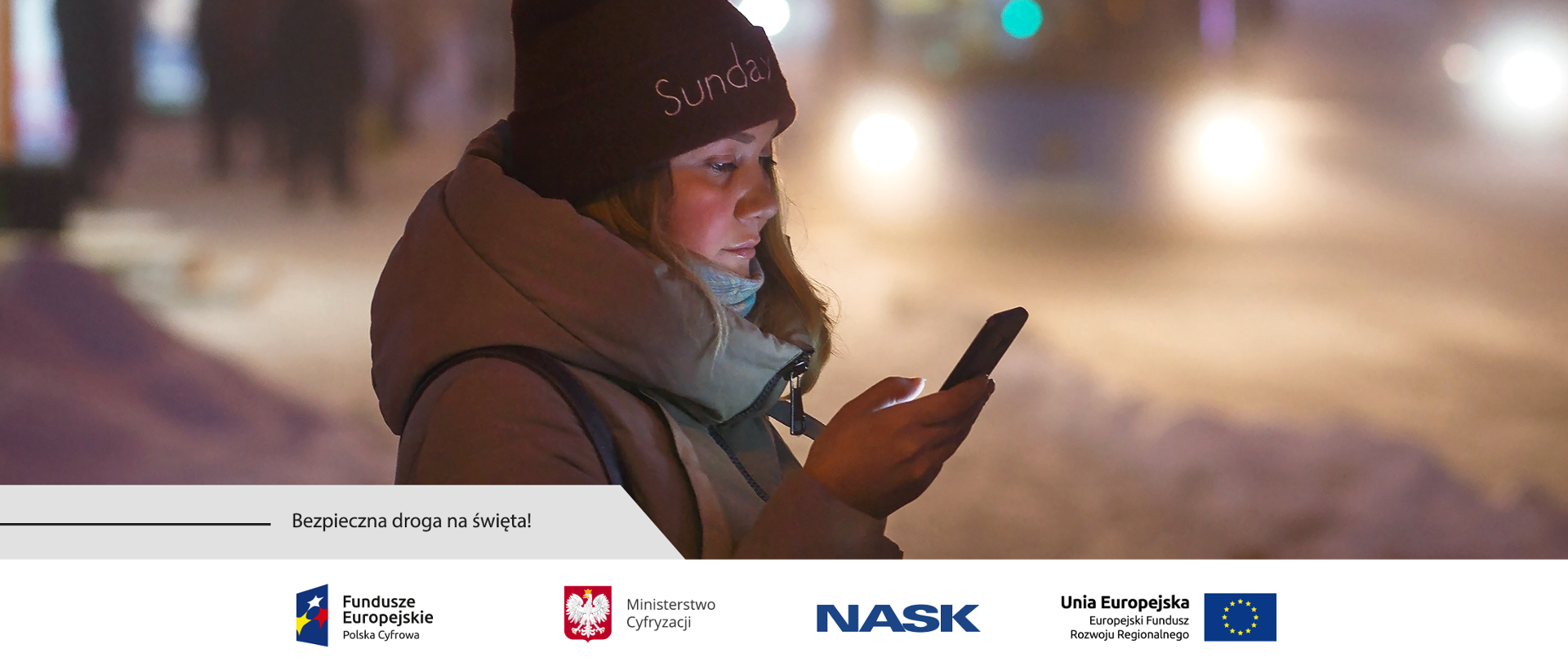 Zimowa aura. Na chodniku stoi dziewczyna trzymająca w prawej dłoni telefon komórkowy. W tle światła nadjeżdżającego pojazdu i ludzie na przystanku autobusowym.