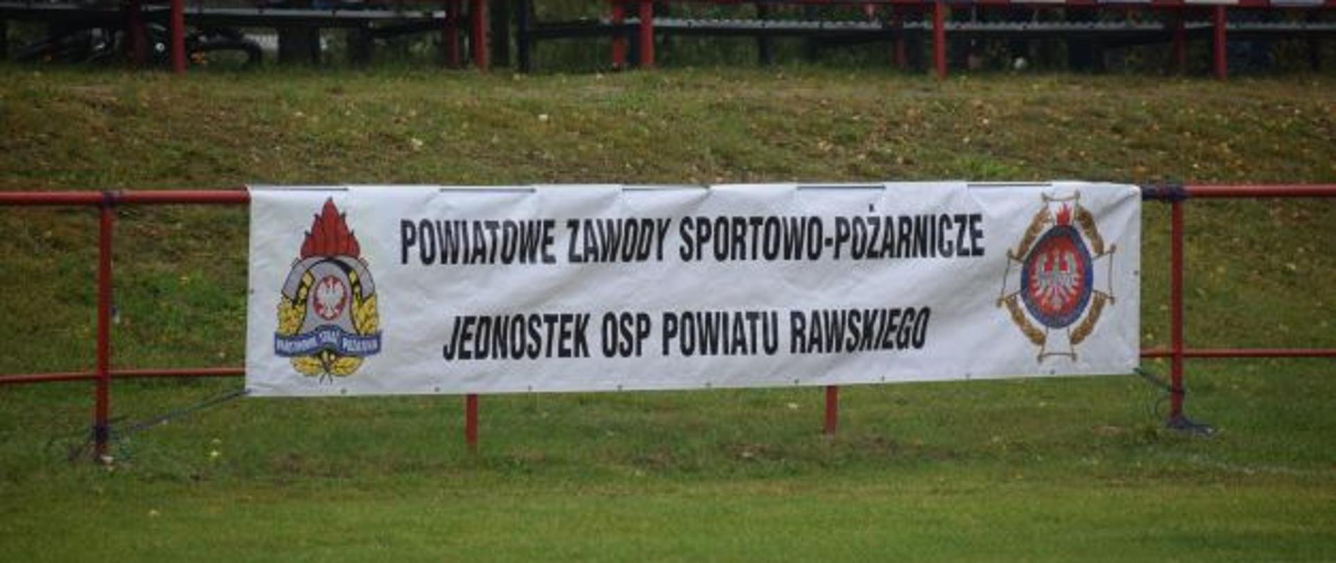 Na ogrodzeniu wisi biały baner z napisem Powiatowe Zawody Sportowo - Pożarnicze Jednostek OSP Powiatu Rawskiego. Z tyłu widać dwa rzędy kolorowych krzesełek trybun boiska.