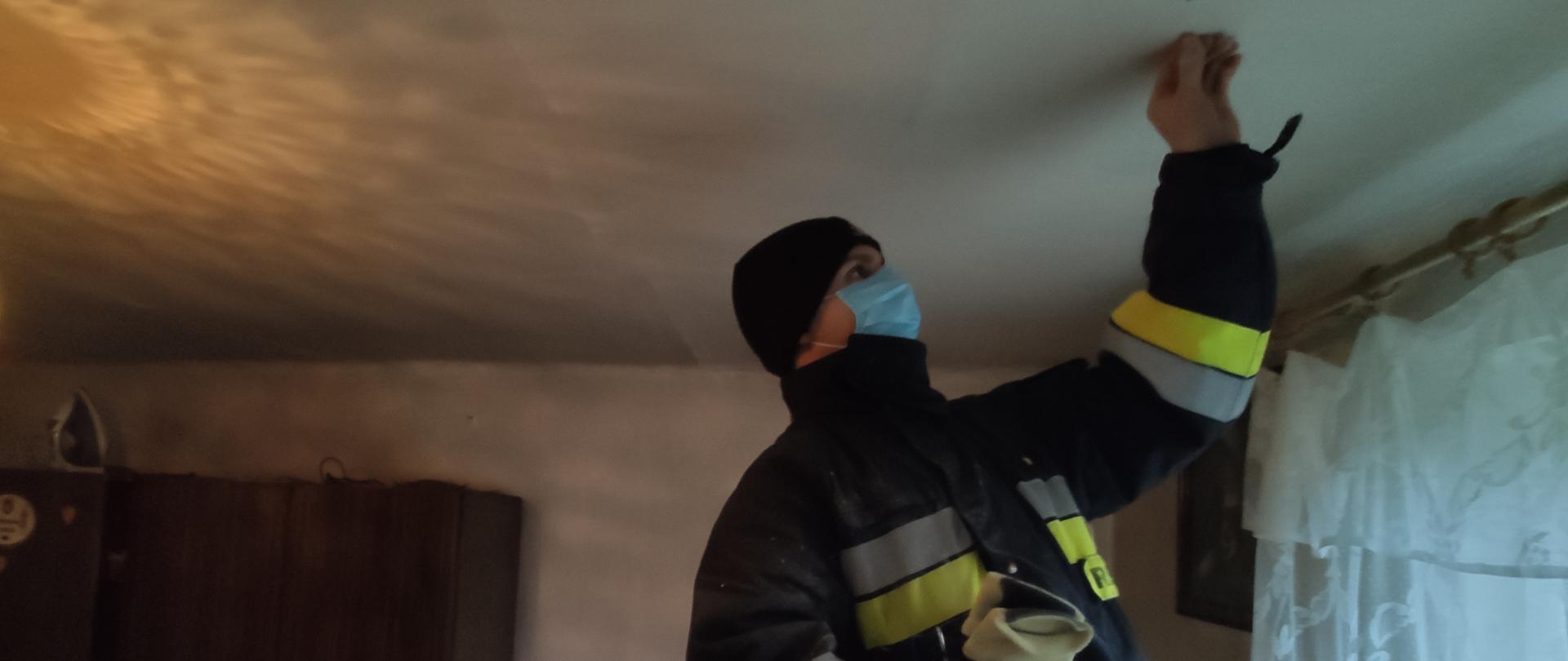 Zdjęcie przedstawia strażaka wykonującego montaż czujki dymu na suficie w pokoju budynku mieszkalnego. W tle umeblowanie pokoju. 