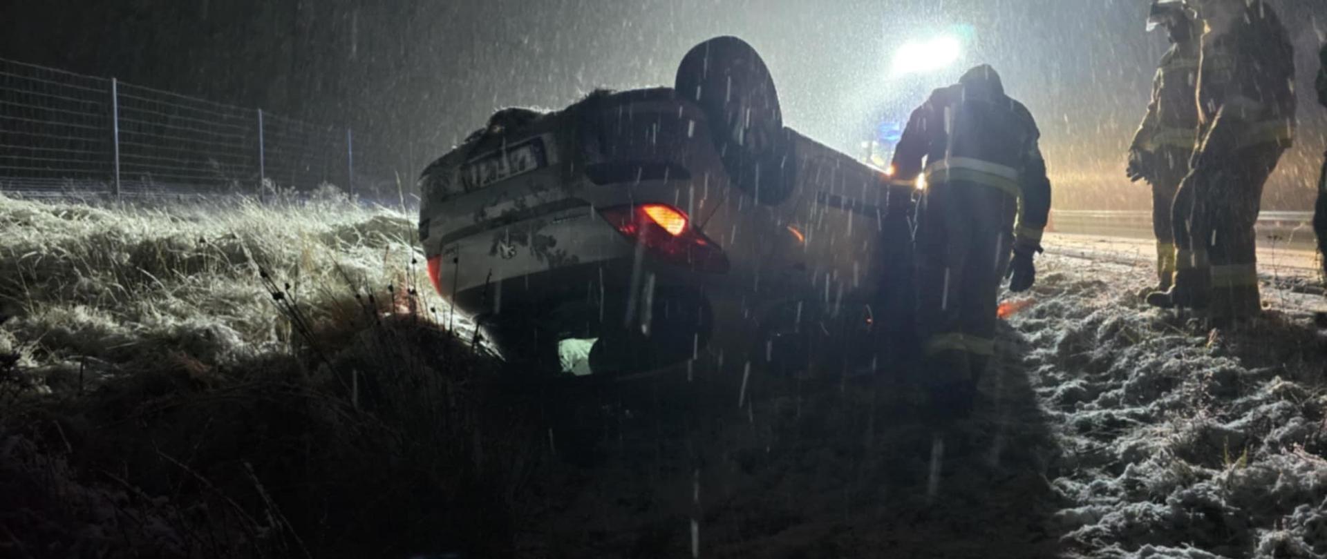 Droga ekspresowa S5. Z lewej strony, w rowie, leży na dachu samochód osobowy. Obok niego znajduje się 3 strażaków uczestniczących w akcji. Z lewej strony płot z siatki odgradzający drogę . Pada śnieg. Na dworze jest ciemno, a teren oświetlany jest przez straż pożarną.