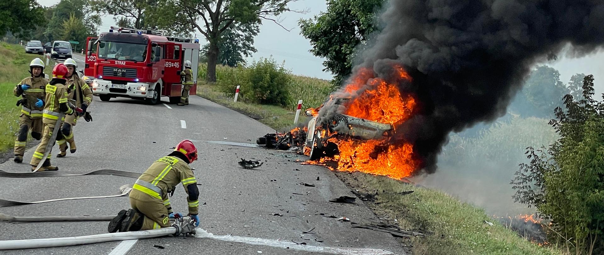 na poboczu drogi pali się samochód przed nim lezy spalony motocykl kawałek przed palącym się samochodem stoi samochód pożarniczy w poprzek drogi jeden strażak klęczy na asfalcie nad rozdzielaczem trzech biegnie do lini wężowej z samochodu wydobywa się czarny dym oraz płomienie dookoła drzewa.
