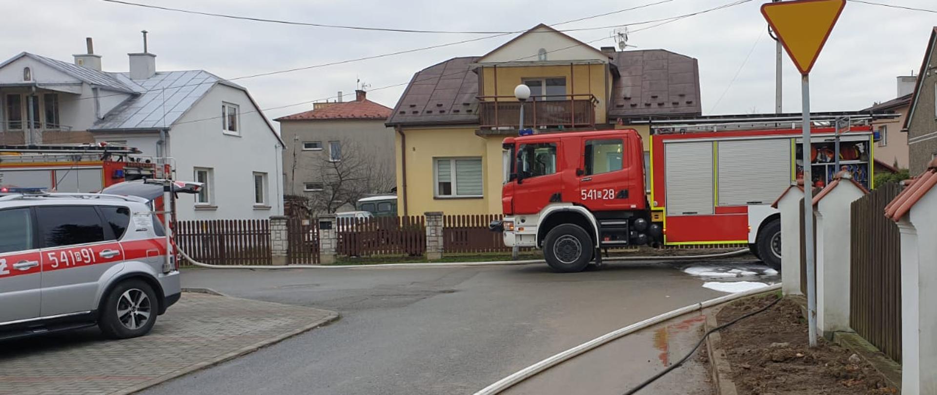 Zdjęcie wykonane na tle budynków mieszkalnych jednorodzinnych w m. Sanok. Na pierwszym planie znajduje się jezdnia, na której ustawione są trzy samochody pożarnicze. Na jezdni znajduje się linia wężowa. 