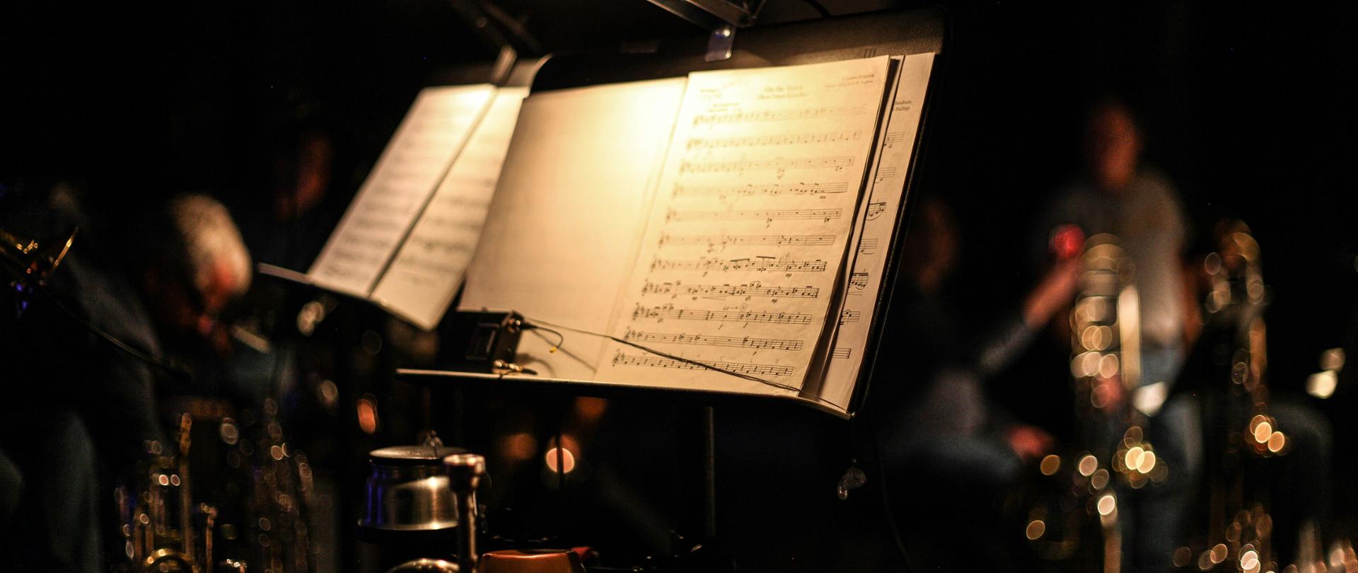 Pulpit nutowy z otwartymi nutami oświetlony dwiema lampkami w ciemnej sali koncertowej. W tle, lekko rozmyci, widoczni są muzycy oraz instrumenty dęte blaszane ustawione na scenie. Całość utrzymana jest w ciepłym, przyciemnionym oświetleniu scenicznym.