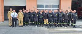 Na zdjęciu widoczna jest grupa strażaków stojących przed budynkiem straży pożarnej, w tle znajdują się bramy garażowe. Część osób ubrana jest w piaskowe mundury, natomiast inni mają na sobie czarne uniformy z elementami odblaskowymi. Strażacy ustawieni są w rzędach, pozując do wspólnego zdjęcia w słoneczny dzień.