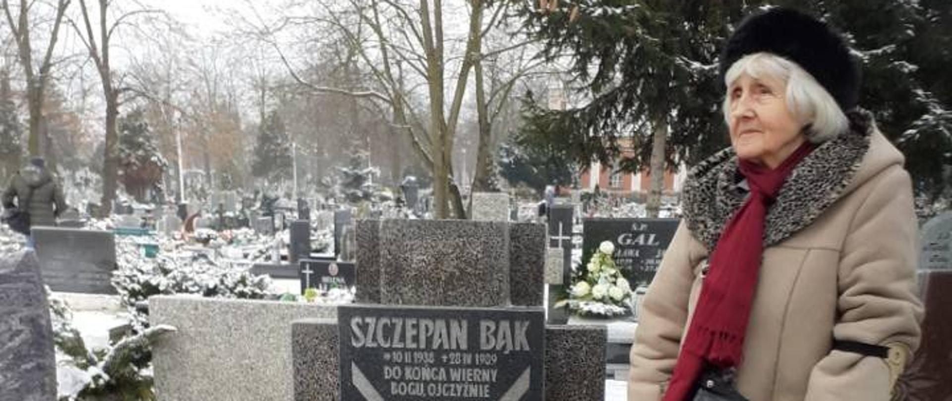 Janina Bąk stoi przy grobie męża, na którym leży biało-czerwony wieniec z szarfami Premiera RP
