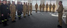 Po lewej stojąca w sali, w szyku grupa strażaków rozpoczynających szkolenie, naprzeciw stojący Zastępca Komendanta Centralnej Szkoły PSP. W głębi stojący przy ścianie okiennej sali funkcjonariusze PSP będący świadkami wydarzenia