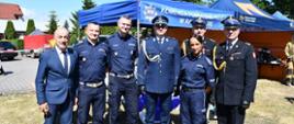 Grupa funkcjonariuszy Policji, strażaków PSP oraz dyrektora szkoły pozuje do wspólnego zdjęcia na terenie pikniku. W tle widoczne są namioty edukacyjne służb mundurowych, w tym stanowisko Ochrony Ludności i Obrony Cywilnej.