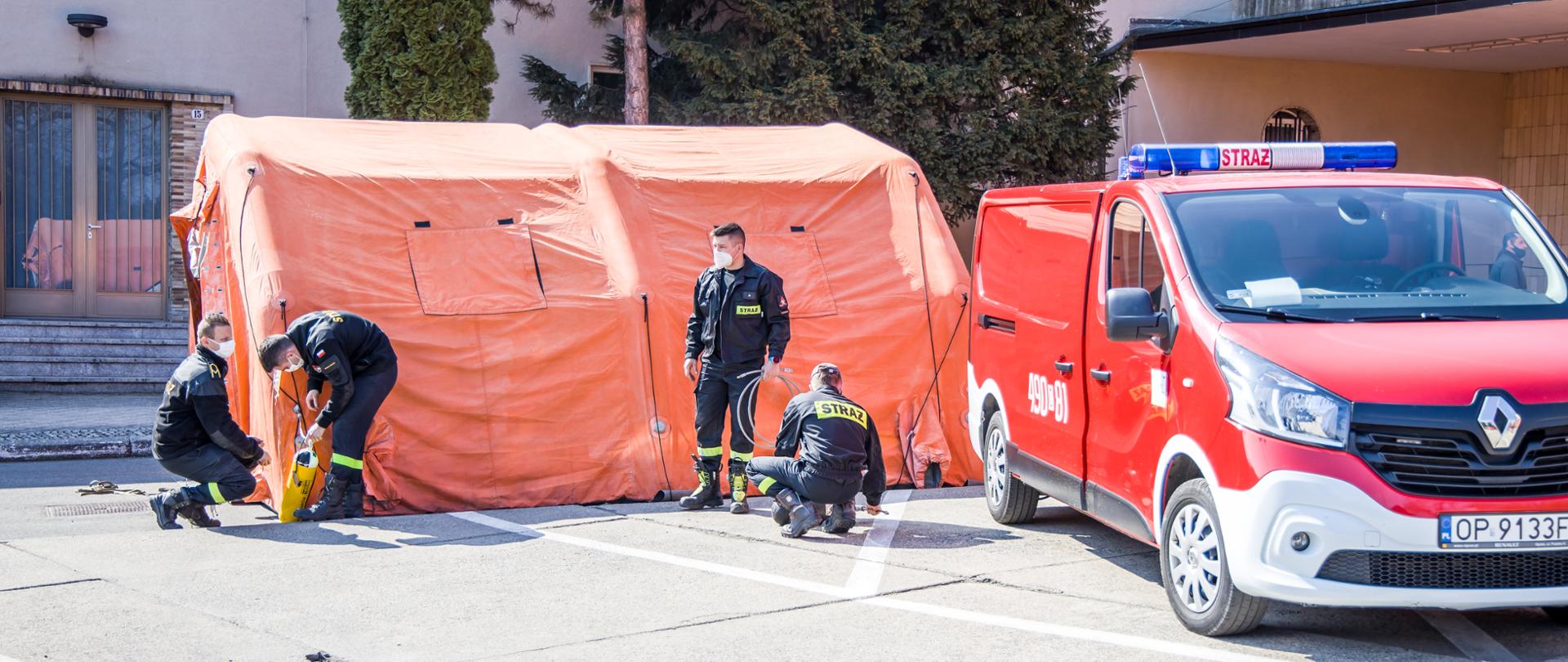 Zdjęcie przedstawia czterech strażaków PSP w umundurowaniu koszarowym podczas sprawiania (pompowania) namiotu pneumatycznego na parkingu Opolskiego Urzędu Wojewódzkiego. Na zdjęciu widoczny samochód kwatermistrzowski koloru czerwonego.