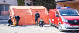 Zdjęcie przedstawia czterech strażaków PSP w umundurowaniu koszarowym podczas sprawiania (pompowania) namiotu pneumatycznego na parkingu Opolskiego Urzędu Wojewódzkiego. Na zdjęciu widoczny samochód kwatermistrzowski koloru czerwonego.