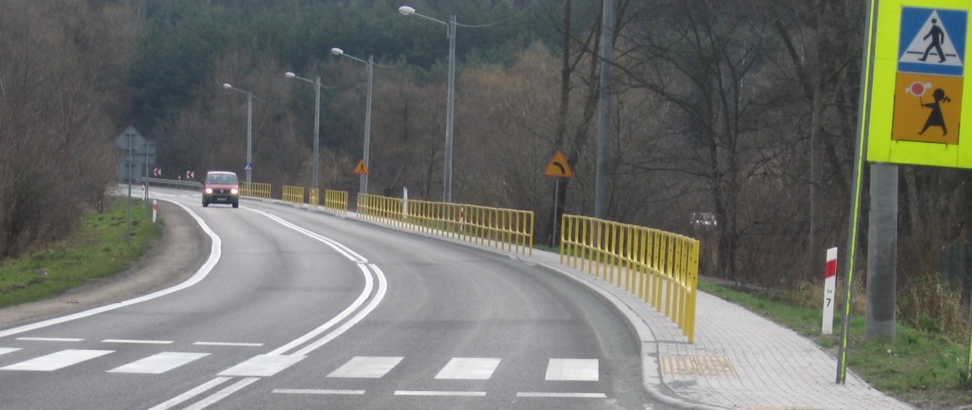 Przejście dla pieszych - jednojezdniowa droga asfaltowa, oznakowane przejście dla pieszych, widoczny zakręt w lewo, z prawej strony drogi chodnik oddzielony od jezdni żółtymi barierami, znaki drogowe i latarnie. Z oddali nadjeżdża drogą czerwony samochód. Przy drodze drzewa i zarośla