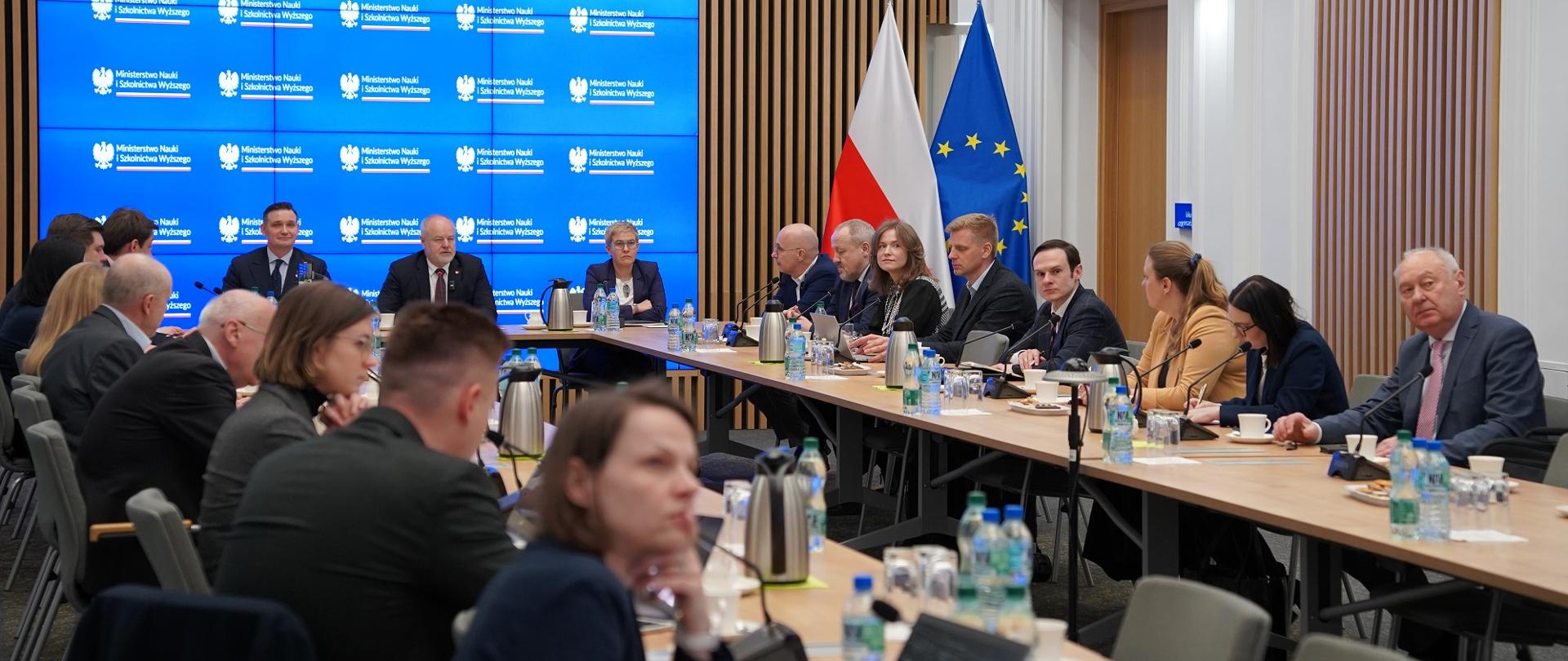 Zdjęcie z tyłu sali pod kątem, za ustawionymi w czworokąt stołami siedzą ludzie, u szczytu stołu wiceminister Gzik, wiceminister Mrówczyńska i mężczyzna w garniturze, za nimi wbudowany w ścianę z pionowych drewnianych listewek wielki monitor.