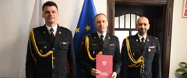 Na zdjęciu w mundurach wyjściowych znajdują się sylwetki trzech komendantów państwowej straży pożarnej. W tle widoczna flaga Polski oraz Unii Europejskiej.