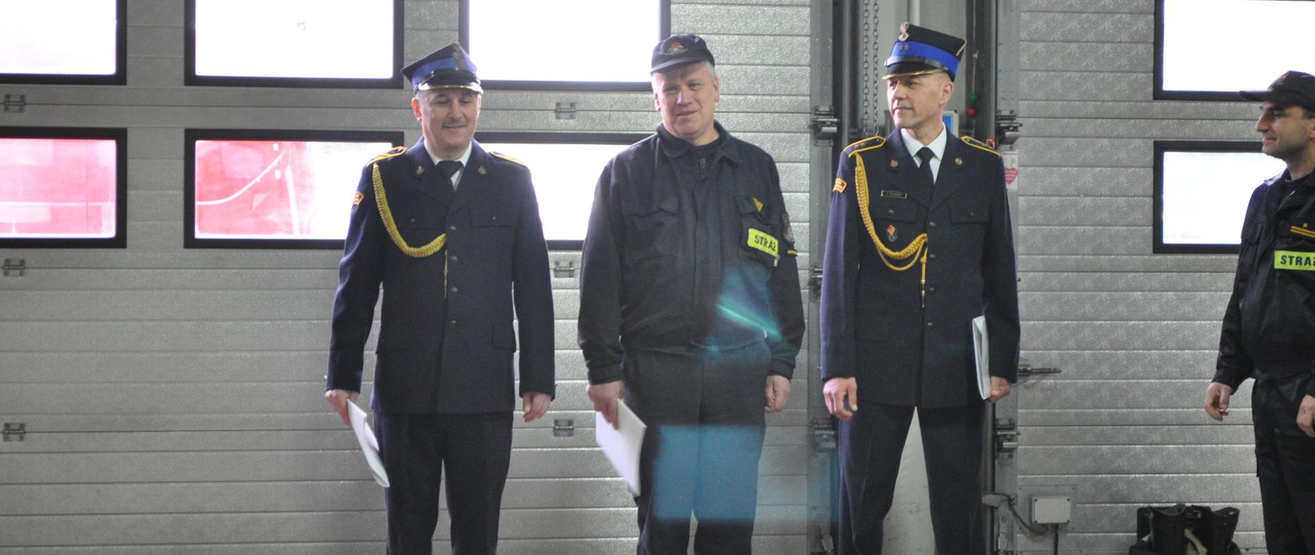 Na zdjęciu w garażu siedziby JRG Nr 1 w Ostrowcu Św. widać strażaków stojących na oficjalnej zmianie służby. Po pierwszym planie zdjęcia widać trzech odchodzących na emeryturę strażaków. W tle bramy garażowe.