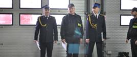 Na zdjęciu w garażu siedziby JRG Nr 1 w Ostrowcu Św. widać strażaków stojących na oficjalnej zmianie służby. Po pierwszym planie zdjęcia widać trzech odchodzących na emeryturę strażaków. W tle bramy garażowe.
