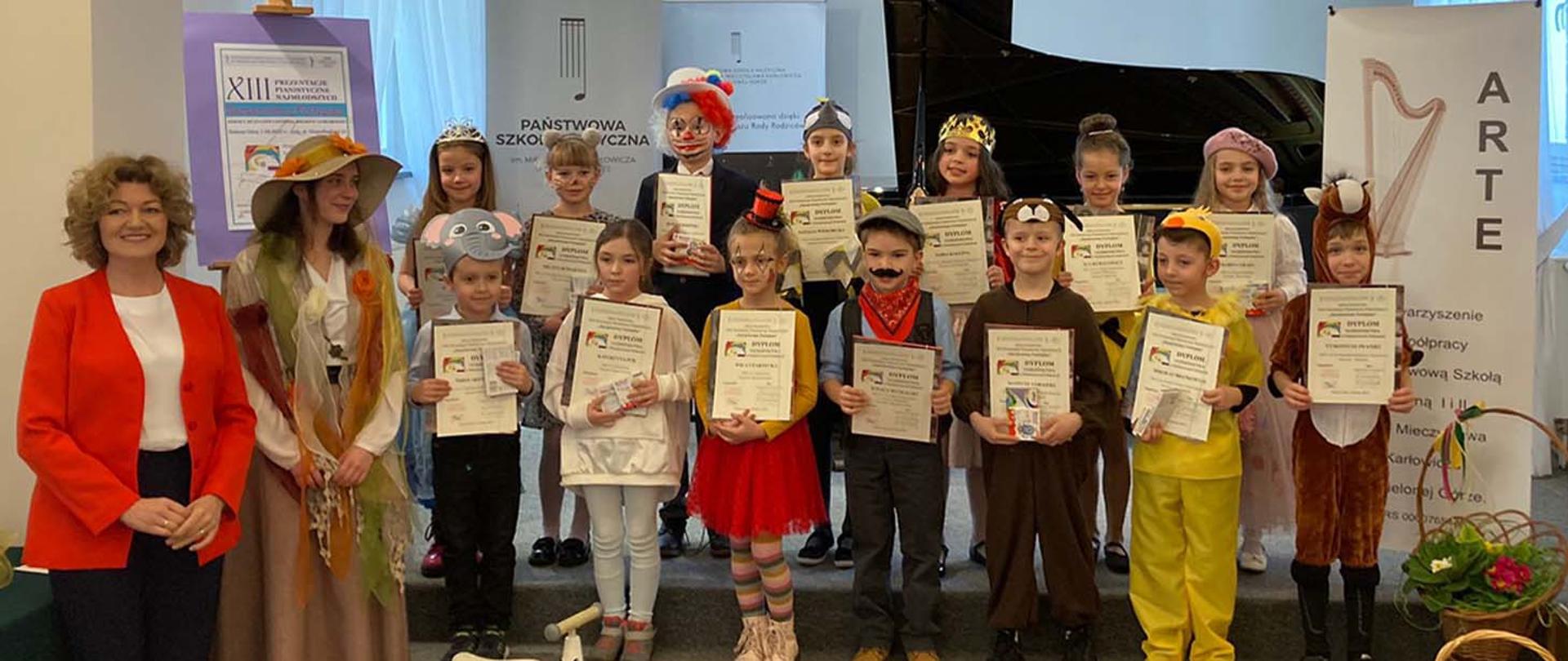 Uczestnicy konkursu fortepianowego pozując do zdjęcia na scenie, dzieci trzymają w rękach dyplomy, obok nich stoją dwie nauczycielki.