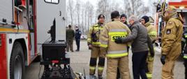 Osoba cywilna stoi ze strażakami Państwowej Straży Pożarnej oraz ochotniczych straży pożarnych stoją obok sprzętu i samochodu specjalnego. 