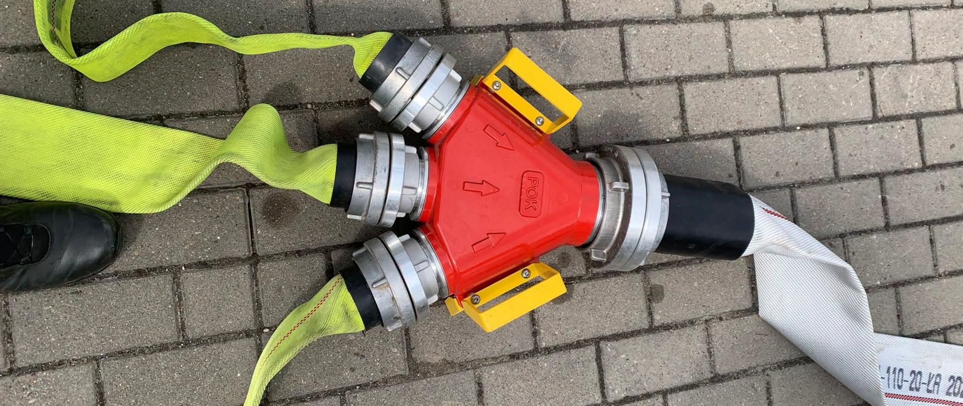 Zdjęcie przedstawia element armatury pożarniczej wykorzystywany podczas ćwiczeń. Łącznik zbierający wodę z trzech węży o średnicy 52 mm i przesyłający ja dalej jednym wężem o średnicy 110 mm