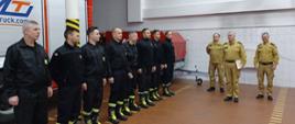 Zdjęcie przedstawia strażaków stojących w rzędzie w pomieszczeniach garażowych jednostki. Po prawej stronie stoi Komendant Powiatowy Państwowej Straży Pożarnej w Człuchowie st. bryg. Radosław Batruch, za nim Zastępca Komendanta Powiatowego PSP w Człuchowie bryg. Henryk Koźlewicz i Dowódca Jednostki Ratowniczo-Gaśniczej w Człuchowie bryg. Piotr Głodowski