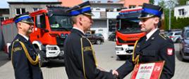 Zdjęcie przedstawia trzech strażaków jeden w reku trzyma dyplom oraz dokonywany jest uścisk ręki w tle samochody pożarnicze