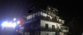 Na zdjęciu widoczny wielokondygnacyjny budynek mieszkalno - usługowy posiadający uszkodzenia elewacji oraz poddasza po ugaszeniu pożaru. Z lewej strony mocne światło z samochodu pożarniczego oświetlające teren działań. 