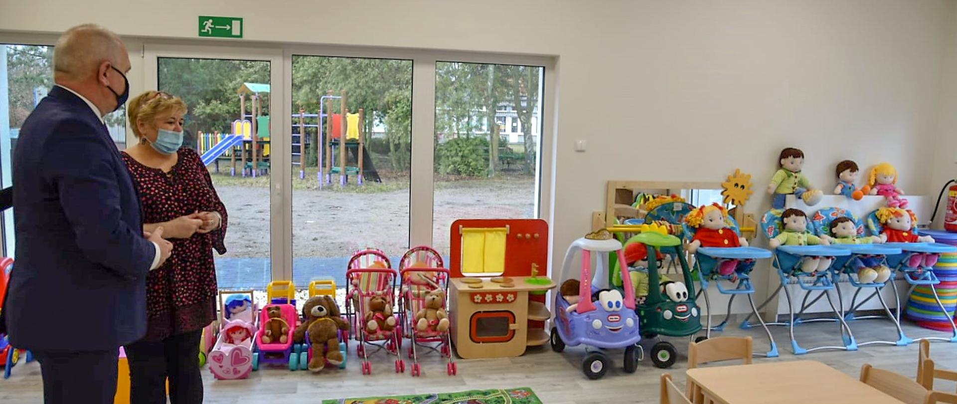 Wojewoda Lubuski rozmawia z dyrektor placówki w jednej z sal. Widok na zabawki i wyposażenie. W tle okna z widokiem na plac zabaw