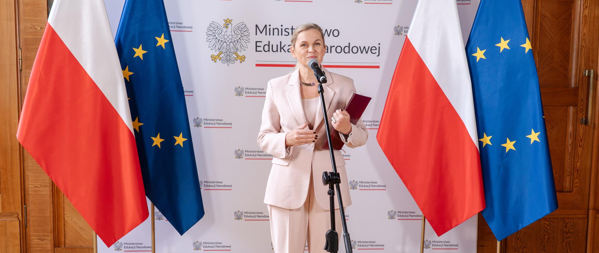 Minister Barbara Nowacka mówi do mikrofonu w czasie konferencji prasowej na której ogłasza nowe programy ministra na 2025 rok. Ubrana w jasnoróżowy garnitur. W ręku trzyma teczkę z orłem