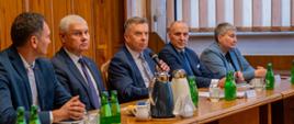 Minister Wieczorek siedzi między czterema mężczyznami w garniturach za drewnianym stołem i mówi do mikrofonu, za nimi wyłożona drewnem ściana.