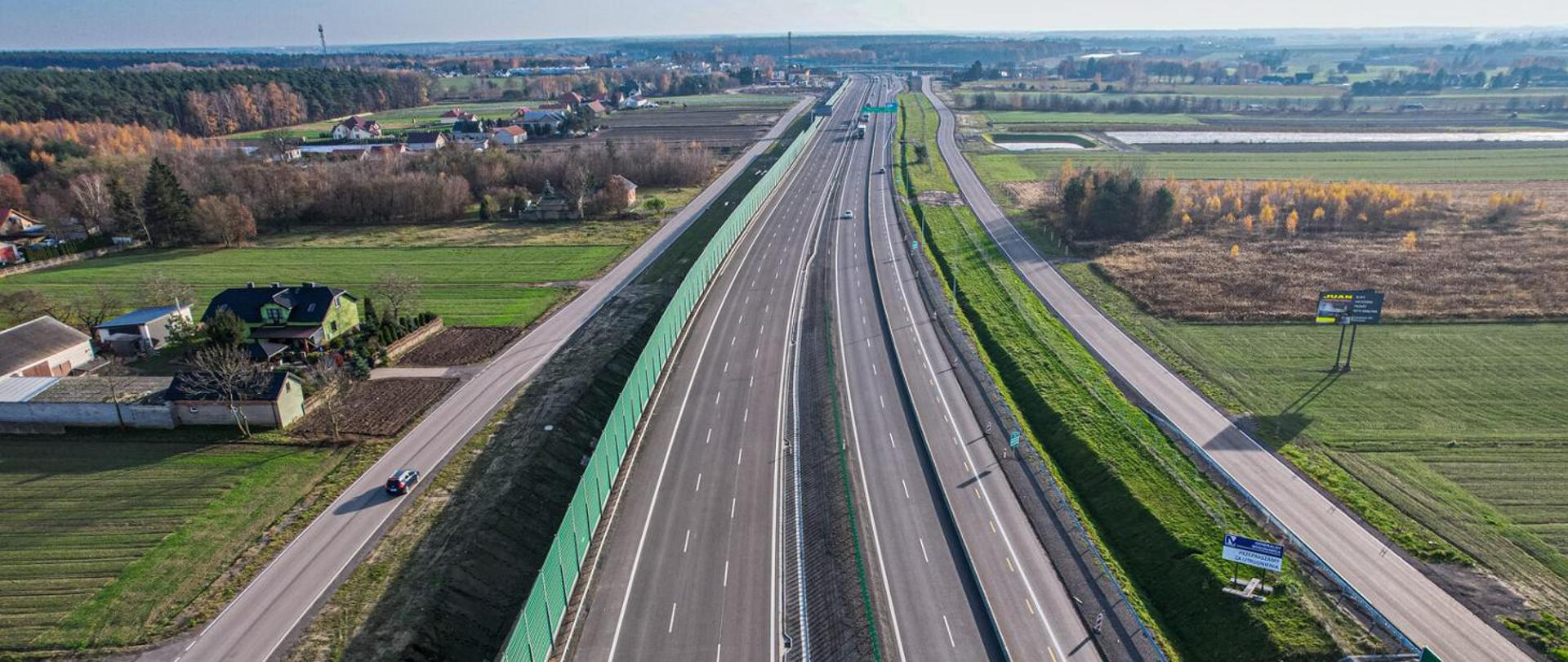 Na zdjęciu widać fragment drogi ekspresowej S7 w rejonie Załuski–Modlin. Droga przebiega przez teren otwarty. W tle widoczne są pola i niska roślinność, a także linia drzew w oddali.
Jezdnia jest asfaltowa, z wyraźnymi pasami ruchu i oznakowaniem poziomym. Po obu stronach drogi znajdują się barierki energochłonne (metalowe).
Widoczny jest pas rozdziału między kierunkami ruchu. Dominują odcienie szarości (asfalt), zieleni (roślinność) i niebieskiego (niebo). Niebo jest częściowo zachmurzone, ale jasne, co sugeruje dobre warunki pogodowe.