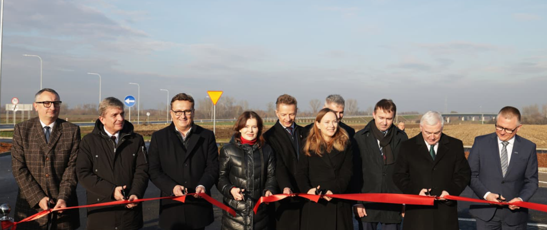 Na zdjęciu widzimy uroczystość otwarcia obwodnicy Nowego Korczyna. Grupa osób stoi przy przecięciu wstęgi, symbolizującym zakończenie inwestycji. Wśród uczestników wydarzenia znajduje się Pani Katarzyna Pełczyńska-Nałęcz, minister funduszy i polityki regionalnej, która wyróżnia się czerwonymi spodniami. Pozostali obecni trzymają nożyczki i przygotowują się do wspólnego przecięcia wstęgi. W tle widać nową drogę i oznakowanie. Jest słoneczny dzień, a pogoda sprzyja uroczystościom.
