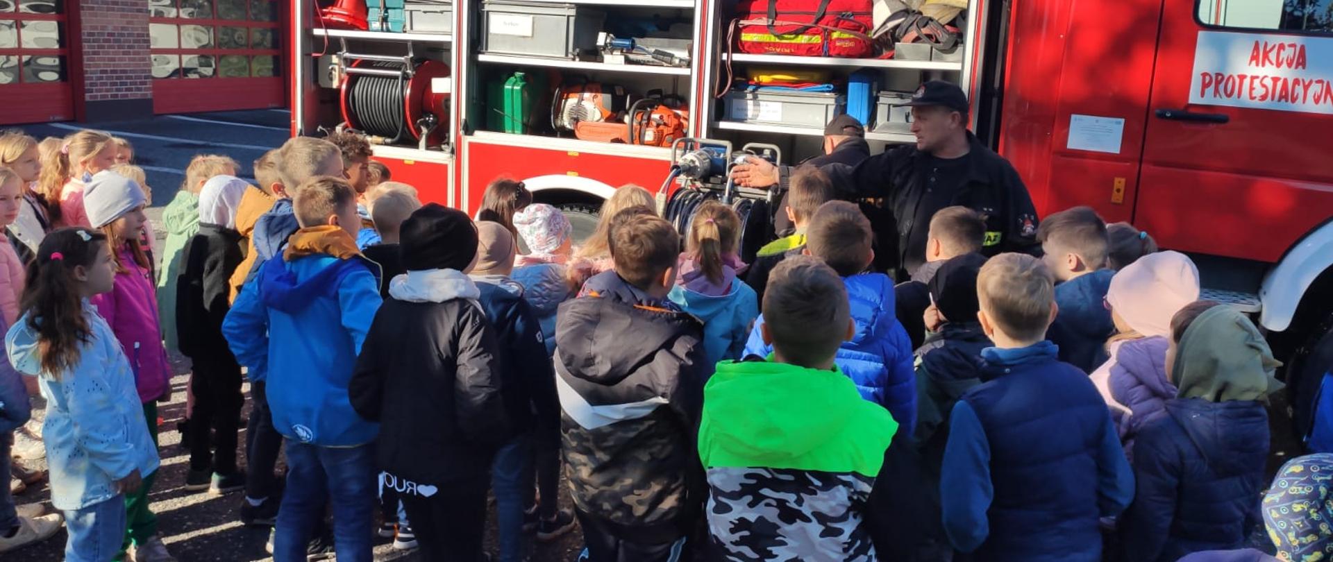 Na zdjęciu znajdują się dzieci oglądające wóz strażacki oraz strażak, który im prezentuje sprzęt pożarniczy