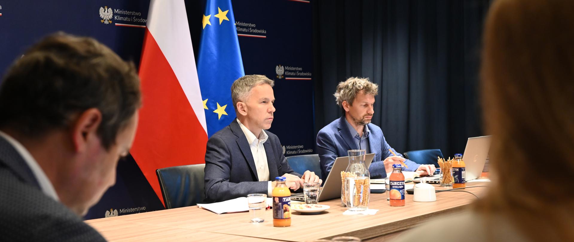 Cztery osoby siedzą z komputerami przy biurku. W centrum na tle flagi polskiej i EU siedzą członkowie Międzyresortowego Zespołu ds. Odry