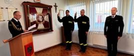 Z prawej strony zdjęcia widzimy dwóch młodych mężczyzn z uniesionymi prawymi rękami do przysięgi ubranych w czarne ubrania koszarowe z jasnożółtymi emblematami, z czarnym napisem "STRAŻ". Po lewej stronie stoi dojrzały mężczyzna ubrany w białą koszulę z czarnym krawatem, marynarką czarną, trzymający w ręku dokumenty. Po jego prawej stronie stoi mównicza z godłem Rzeczypospolitej Polskiej. W tle widzimy Sztandar KP PSP Świdwin, Krzyż. 