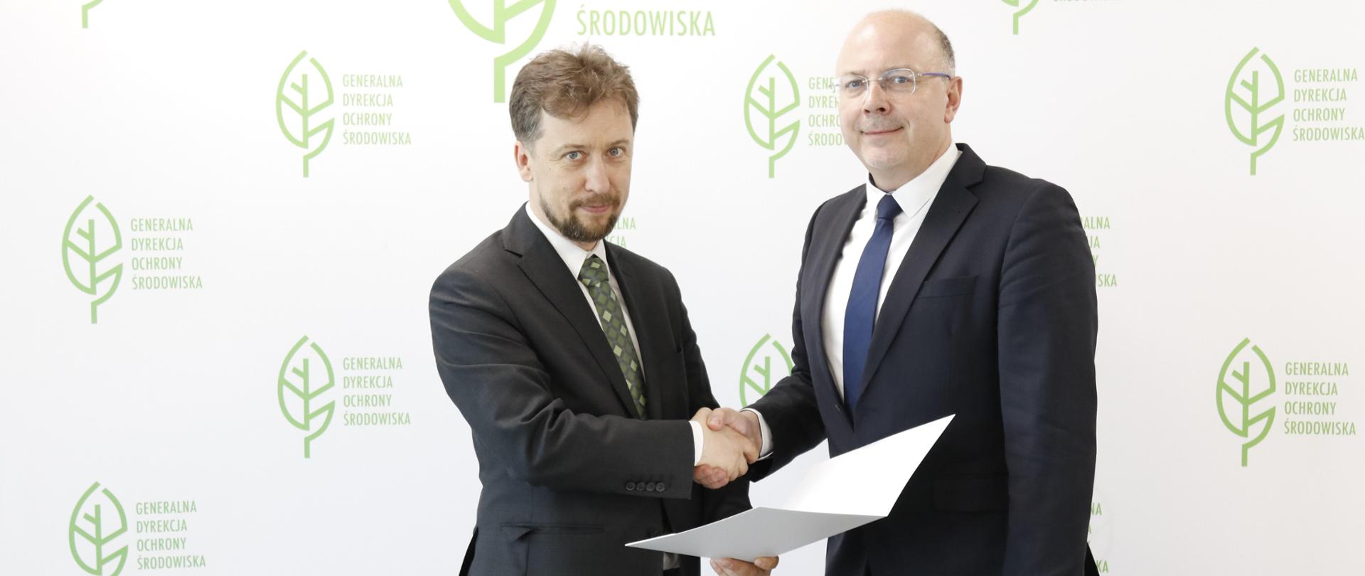 Na tle białej ścianki z logotypami Generalnej Dyrekcji Ochrony Środowiska (zielony liść) stoją dwaj elegancko ubrani mężczyźni. Ściskają sobie dłonie. Jeden trzyma otwartą teczkę. 