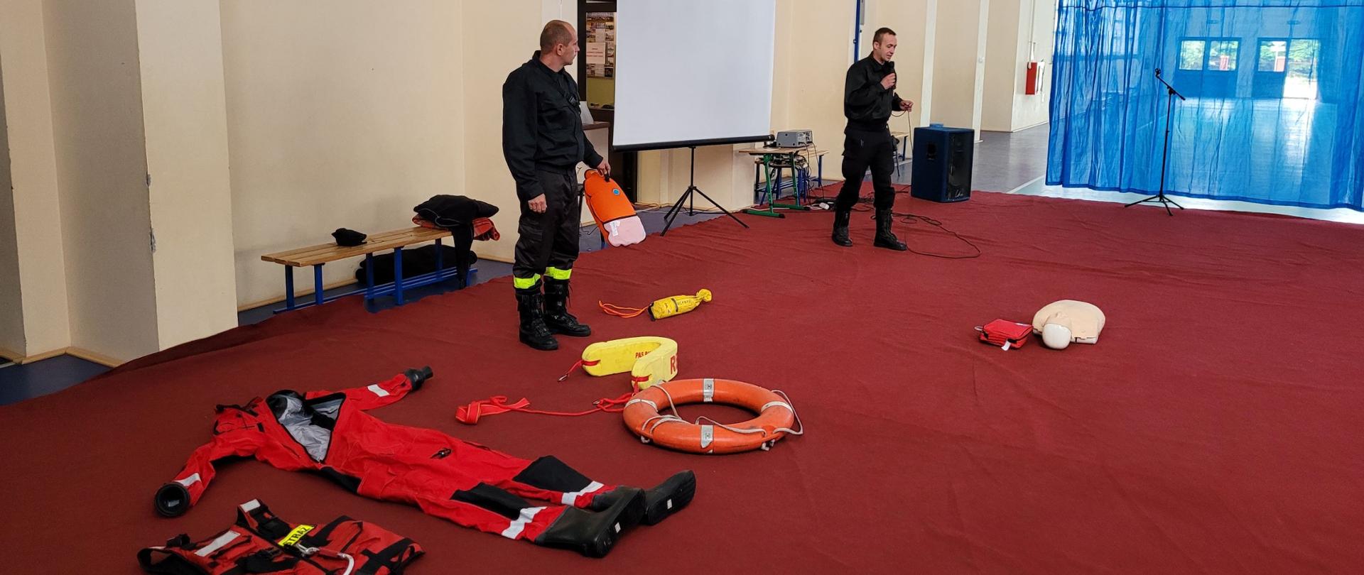 Zdjęcie przedstawia dwóch mężczyzn w czarnych mundurach w dużym pomieszczeniu z czerwoną wykładziną na podłodze, którzy prowadzą spotkanie. Przed nimi leżą czerwone koło ratunkowe, kamizelka asekuracyjna i kombinezon. W tle kremowe ściany pomieszczenia i niebieska siatka oddzielająca..