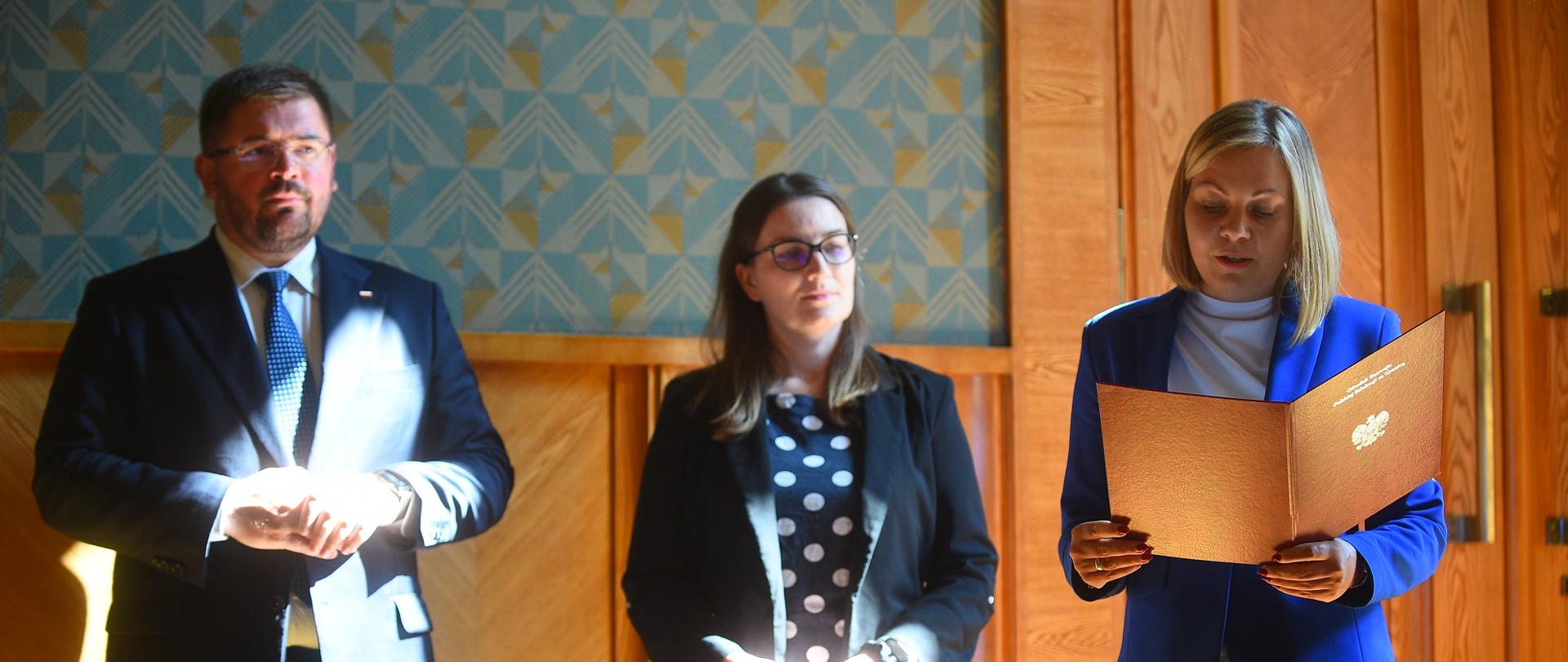 Pod zieloną ścianą stoi wiceminister Rzymkowski i dwie kobiety, jedna czyta z otwartej teczki.